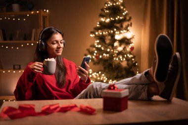 Mutlu kadın Noel günü akıllı telefon ve kulaklık kullanarak müzik dinliyor. Boşluğu kopyala