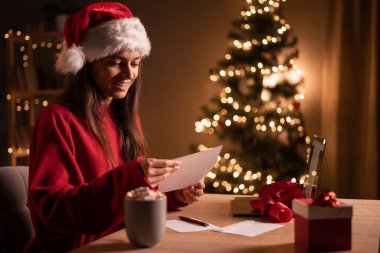 Evdeki masada oturan ve elinde Noel kartı olan bir zarf tutan genç bir kadın. Noel dilekleri, tatiller ve kutlama konsepti. Boşluğu kopyala