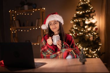 Genç bir kadının portresi dizüstü bilgisayara sarılı battaniyeye sarılı oturma odasında Noel Baba şapkası takıyor. Boşluğu kopyala