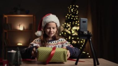 Mutlu Xmas kızı evden video görüşmesi yapıyor, Noel ağacındaki masada akıllı telefonu kullanıyor, hediye kutusunu tutuyor, hediyeleri açıyor. Genç kız, blogcu yeni yıl kutlaması ilanı kaydediyor. Boşluğu kopyala