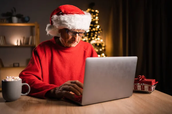 Noel Baba Şapkalı Yaşlı Adam Evde Noel Arifesinde Dizüstü Bilgisayarından — Stok fotoğraf