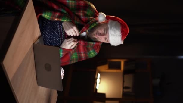照片上的帅哥坐在桌子上拿着笔记本电脑吃棉花糖喝可可 穿着格子呢在装饰过的圣诞客厅里 垂直录像 — 图库视频影像