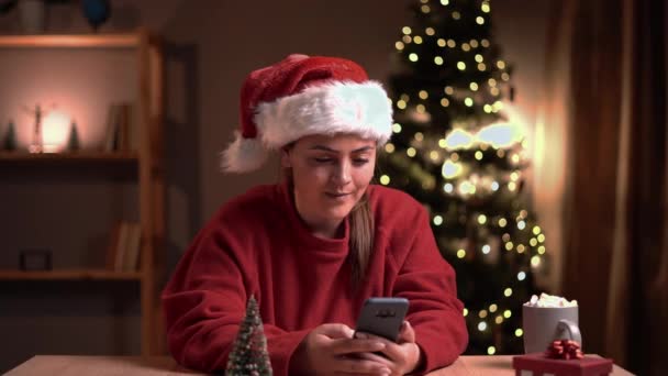 驚きの顔を持つスマートフォンを使用してクリスマスツリーの近くの居心地の良い部屋に座ってサンタの帽子の喜び白人女性はクリスマス宝くじに勝つ 明けましておめでとうございます メリークリスマスのコンセプト — ストック動画