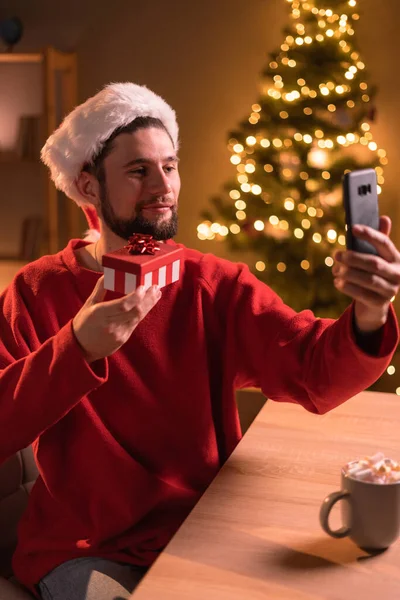 携帯電話を介してビデオ通話を持っている若い幸せな男と自宅でクリスマスを祝いながら カメラでギフトボックスを表示します スペースのコピー — ストック写真