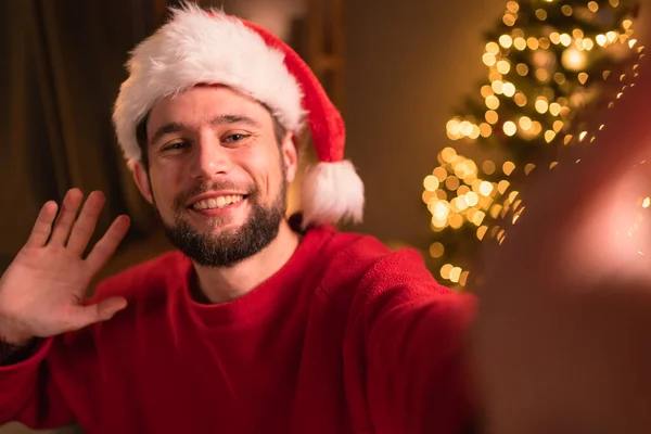 サンタクロースの帽子の幸せなひげを生やした男は 携帯電話を手に手を振って自撮り ビデオ通話 録画ビデオメリークリスマスの挨拶やVlogを撮影します — ストック写真