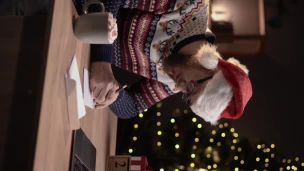 クリスマスの間に自宅のオフィスで赤い帽子の挨拶カードや手紙を書いて身に着けている男性 垂直ビデオ — ストック動画