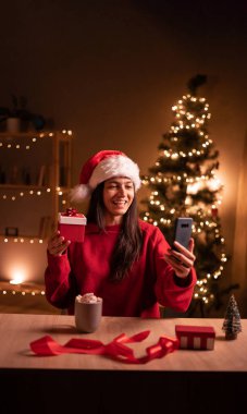 Mutlu İspanyol kadın Noel arifesinde evde akıllı telefondan video çekerken hediye kutusu gösteriyor. Boşluğu kopyala