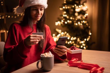 Heyecanlı İspanyol kadın Noel hediyesi sipariş ediyor, rahat bir odada cep telefonu ve kredi kartı kullanıyor. Boşluğu kopyala