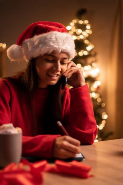 Akıllı telefonlu genç bir kadın Noel tatilinde online alışveriş yapıyor, cep telefonuyla konuşan gülümseyen bir kadın, ev ofisinde çalışırken notlar yazıyor. Boşluğu kopyala