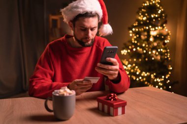Elinde kredi kartı tutan yakışıklı sakallı bir adam ve Noel tatilinde akıllı telefondan ödeme yapıyor. Yılbaşı ağacı ile evde otururken internetten hediyeler alıyor. Boşluğu kopyala