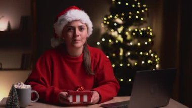 Mutlu arkadaş canlısı kadın elinde bir hediye kutusu tutarken dizüstü bilgisayar kullanıyor ve Noel akşamı kameraya gösteriyor. Boşluğu kopyala