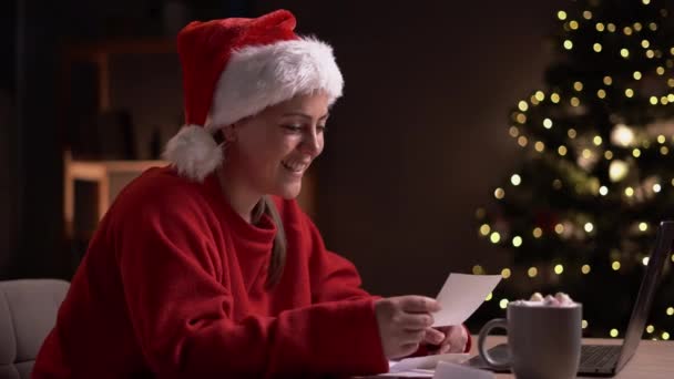 セーターとサンタ帽子を身に着けている幸せな若い女性は クリスマスの手紙を開き 夕方遅くに自宅のオフィスで働いて 家族からグリーティングカードを読んでいます スペースのコピー — ストック動画
