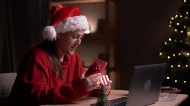 Genç, mutlu bir kadın Noel hediyesini açarken akşam evdeki bilgisayardan bir video görüşmesi yapıyor. Boşluğu kopyala