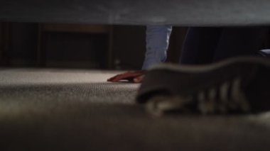 Adam ayakkabılarını kaybetmiş, ayakkabılarını kanepenin altında arıyor. Yakın plan.