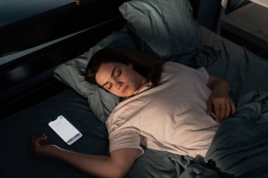 Mobil bağımlılık konsepti. Sosyal medyaya bağımlı genç bir kadın gece yatakta ekranı açık akıllı cep telefonuyla uyuyakalıyor. Karanlık yatak odasında uyuyan kız. Üst görünüm