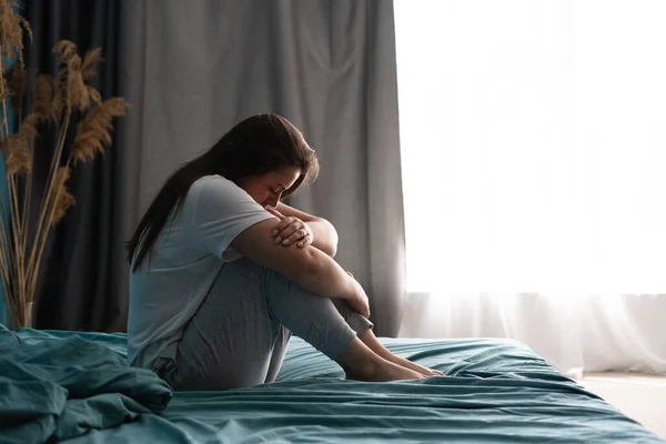 stock image Sad woman sitting on bed in bedroom with light from window. Abuse concept. Copy space