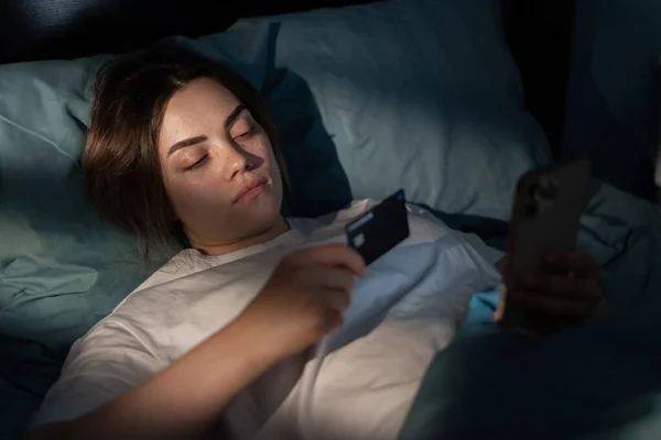 stock image Attractive woman with credit card and smartphone lying in bed at night, looks at the phone screen, shopping online. Young girl awake late at night using smart phone in a dark bedroom. Copy space