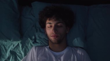 Geceleri yatak odasındaki bir yatakta rahat bir şekilde uyuyan yakışıklı genç adam manzarası. Pencereden süzülen mavi ışık. Yakın plan.
