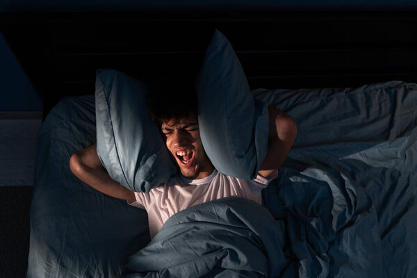 Irritated young man laying in bed at night, covering his head with pillows, stress from noise, sleepless guy having troubles with sleeping. Noisy neighbours and insomnia concept. Top view