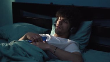 Genç dingin bir adam, karanlık bir odada uyurken kulaklıkla müzik dinlerken akıllı telefon kullanıyor. Boşluğu kopyala