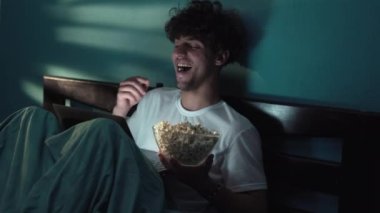 Mutlu gülümseyen genç adam yatakta yatarken patlamış mısır yiyor ve geceleri dizüstü bilgisayarında komedi filmi izliyor. Boşluğu kopyala