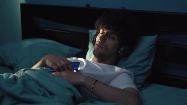 Jonge Serene Man Slapen Bed Donkere Kamer Tijdens Het Luisteren — Stockvideo