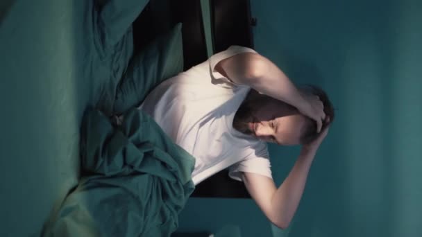 ベッドに座っている落ち込んでいる男は不眠症から眠ることができず 夜に頭痛に悩まされる 垂直ビデオ — ストック動画