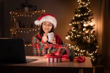 Battaniyeye sarılı genç bir kadın oturmuş film seyrediyor dizüstü bilgisayar kullanıyor, kahve fincanı tutuyor ve dekore edilmiş Noel salonundaki ekrana bakıyor. Boşluğu kopyala