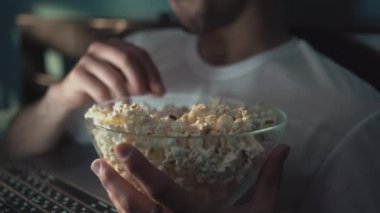 Bekar bir adam gecenin bir yarısı online televizyon izliyor. Yatak odasındaki bir yatakta oturuyor. Patlamış mısır yiyorum. Yakın plan.