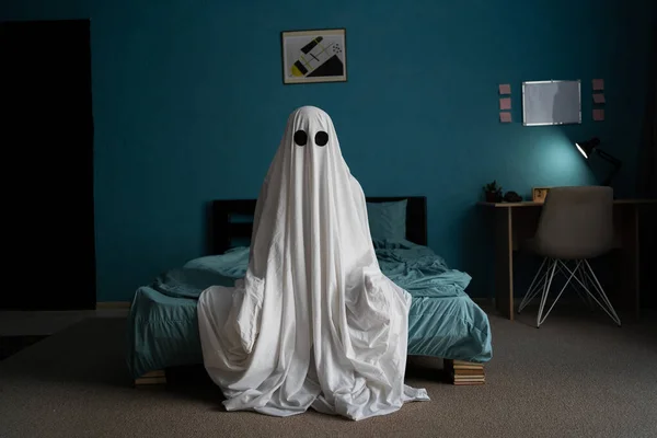 Ghost in white sheet sits on bed in bedroom at night. Horror scene. Halloween concept. Copy space