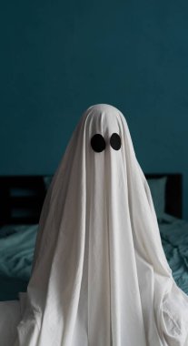 Ghost evdeki yatakta oturuyor, yakın plan. Cadılar Bayramı konsepti Beyaz çarşaflı bir adam. Boşluğu kopyala