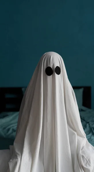 Ghost sitting on the bed at house, close-up. Halloween concept. Man in a white sheet. Copy space