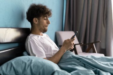 Yakışıklı genç adam, sabah uyandıktan sonra yatağında telefon kullanıyor.