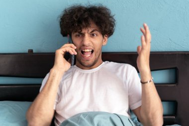 Genç Arap yatakta oturmuş cep telefonuyla konuşuyor ve telefona doğru yüksek sesle bağırıyor, sinirsel stres. Boşluğu kopyala
