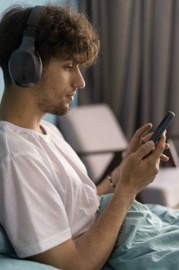 Boş zamanların teknolojisi. Genç Arap adam yatak odasında kablosuz kulaklıkla dinleniyor yatakta akıllı telefon kullanıyor, müzik dinliyor. Boşluğu kopyala