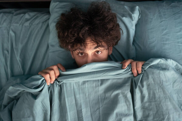Handsome surprised guy covering half of face with blanket, scared man peeking from duvet, afraid of night monsters, top view. Close-up