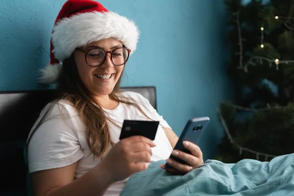 안경을 크리스마스 모자를 침대에 인터넷상에서 선물을 스마트폰으로 카드로 온라인 결제를 — 스톡 사진