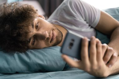 Telefonu kullanan yakışıklı genç adam, kapat, yatakta uzan, sosyal ağda sohbet et, yumuşak yastığa uzan, akıllı telefonu tut ve mesaj yaz..