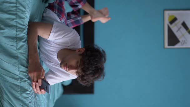 ベッドに寝そべっている若い男のYawnsは スマートフォンが睡眠不足と不眠症 モバイルガジェットへの中毒に苦しんで使用します 垂直ビデオ — ストック動画