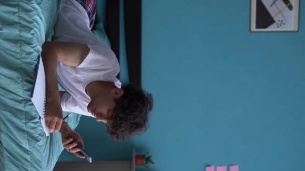 ベッドで勉強している幸せな若い東の男 スマートフォンを使用して インターネットでコースを見て メモを取り パジャマを着て 垂直ビデオ — ストック動画
