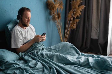 Gülümseyen genç sakallı adam, sabahları yatakta uzanıyor, cep telefonu ve kulaklık kullanarak müzik dinliyor. Boşluğu kopyala