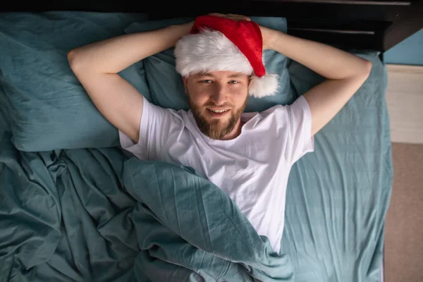 超興奮した男がベッドでサンタの帽子をかぶって幸せを叫んで目を覚ますとクリスマスを祝う準備ができています トップ表示 — ストック写真