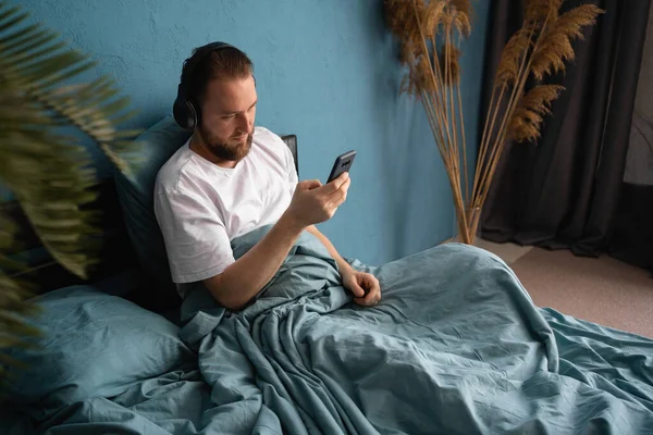 Homme Barbu Couché Sur Lit Dans Casque Regarde Smartphone Écouter — Photo