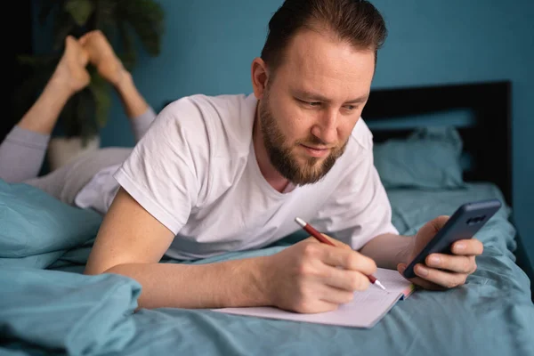 迷人的男人躺在家里的床上 用手机在笔记本上写着便条 后续行动 — 图库照片
