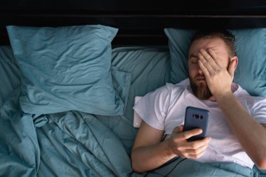 Kafası karışmış bir adam, sabahları evde yatarken cep telefonuna bakıyor. Uyuya kalma konsepti. Üst görünüm