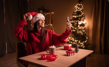 Genç ve mutlu bir kadın Noel arifesinde evde akıllı telefondan video görüşmesi yapıyor. Boşluğu kopyala