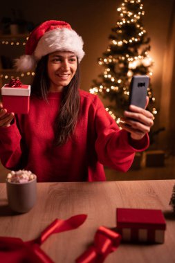 Noel Baba şapkalı neşeli genç bir kadın masada Xmas hediyesi ve akıllı telefonuyla oturuyor, video görüşmesi yapıyor, mobil alışveriş uygulaması kullanıyor, hediyeler alıyor, ailesini ya da evden arkadaşlarını selamlıyor. Boşluğu kopyala