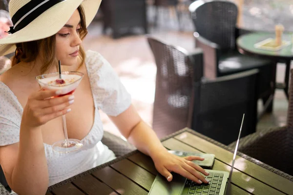 夏のカフェでコンピュータと仕事をわら帽子のビジネス女性 フリーランスのコンセプト ラップトップを使った可愛い若い女性と カフェでストロベリーカクテルを飲んでいます スペースのコピー — ストック写真