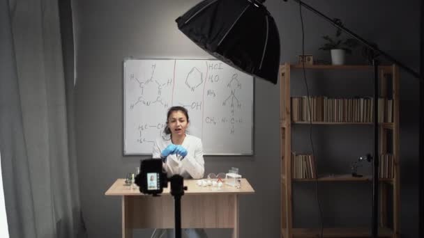 オンライン家庭教師化学教師は自宅のビデオスタジオでVlogを記録します 白衣を着た白人女性は看板の裏側に座っています スペースのコピー — ストック動画