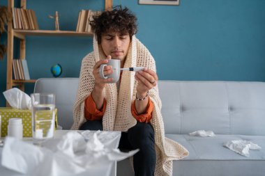 Orta Doğulu hasta bir adam ekose kumaşla kaplanmış kanepede oturmuş sıcak çay içiyor ve elinde termometre tutuyor genç hasta Arap adamın vücut sıcaklığı yüksek, grip belirtileri var. Boşluğu kopyala